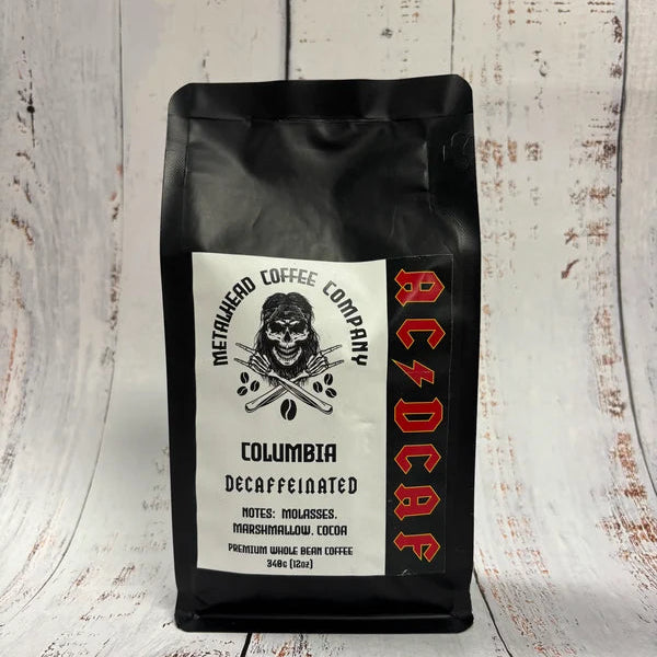 A black bag of Metallica Coffee Company Columbia Dcaffeinated coffee stands on a whitewashed wooden surface with a matching wood plank background. The bag features a white label with a black skull and crossed guitars logo, and the band name 'AC/DC' printed vertically in red on the right side.