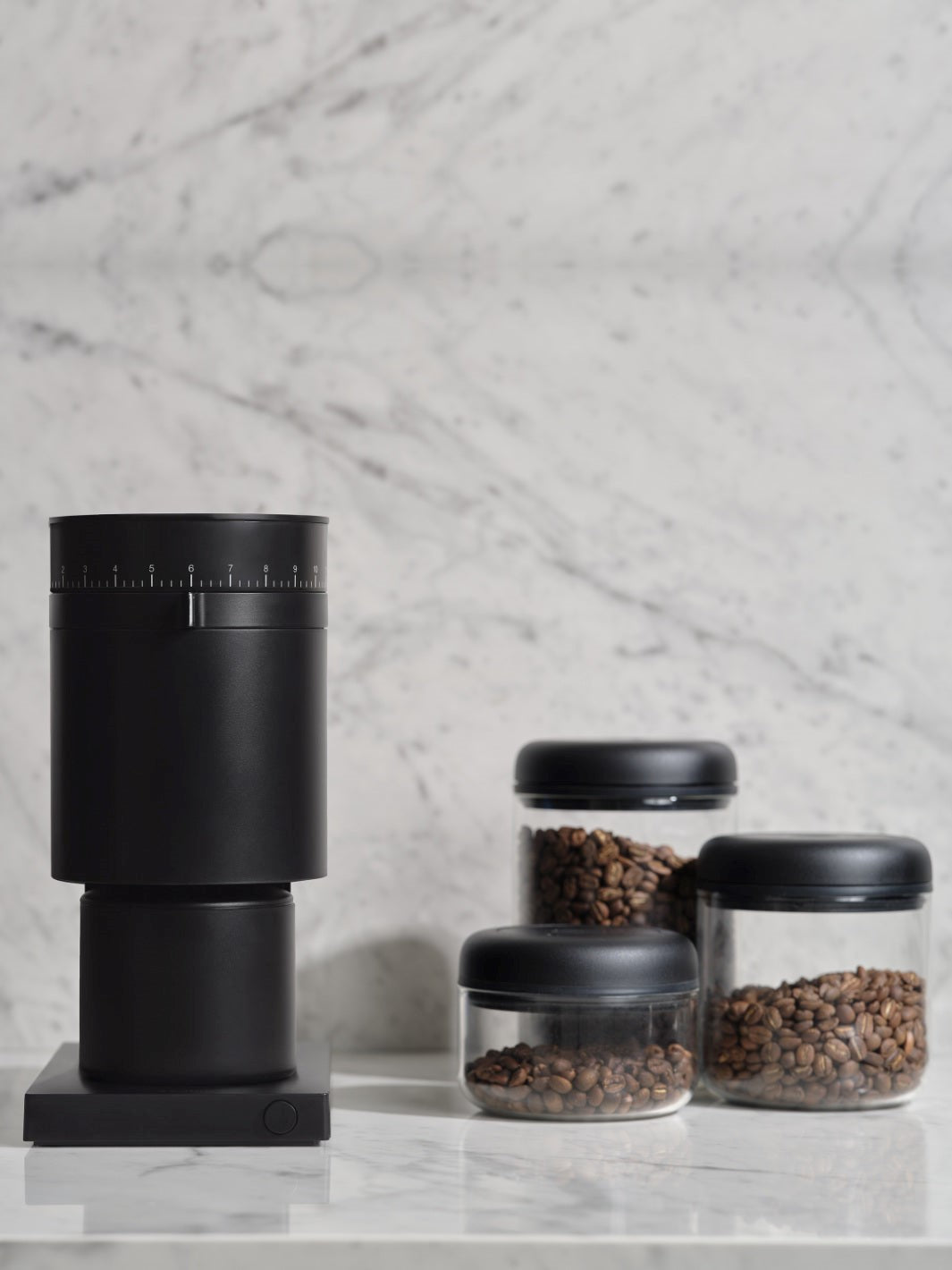 Black coffee grinder with glass Fellow Atmos canisters with coffee beans inside, on a marble surface
