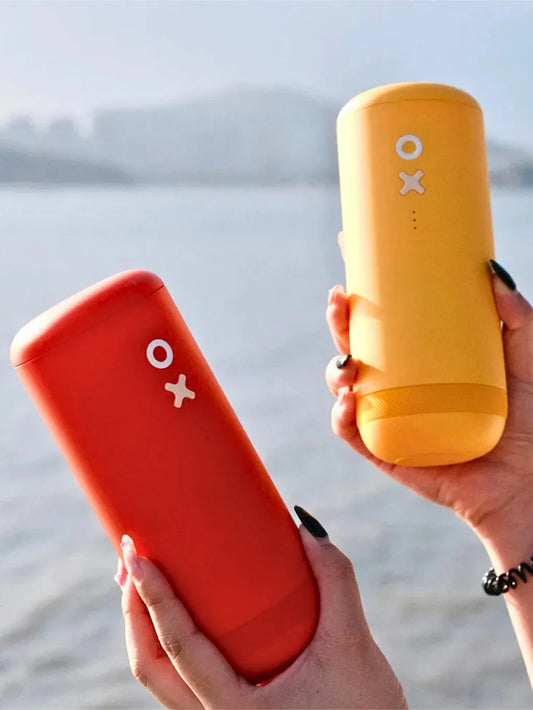 Two portable cylindrical coffee grinders, one orange and one yellow, held in two hands against a blurred outdoor water and mountain background. Both grinders feature a white circular 'o' and cross 'x' logo on the front.