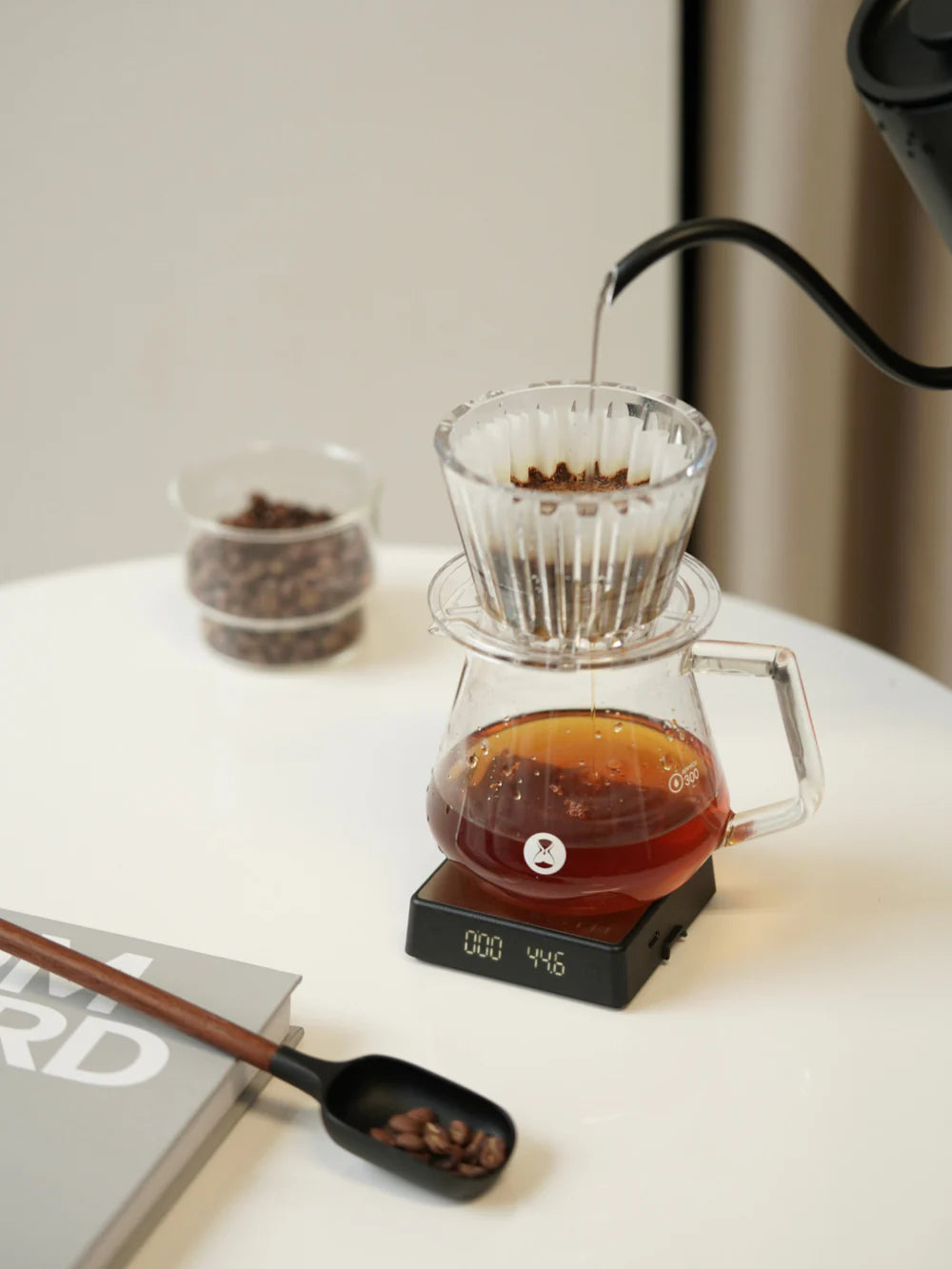Coffee being brewed into a glass carafe on a digital scale with coffee beans and a scoop nearby.
