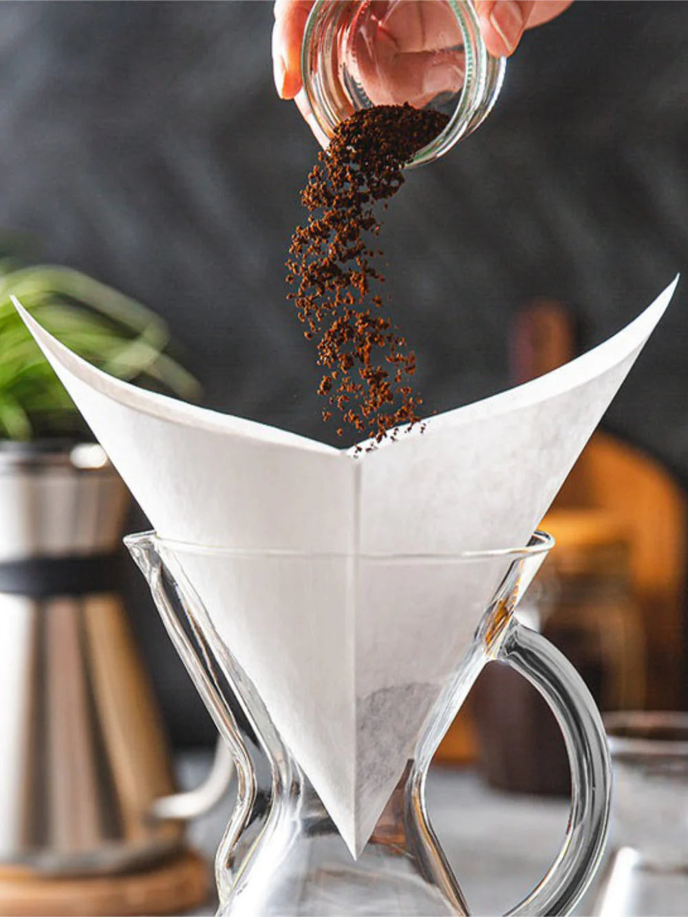 Person pouring coffee grounds into a white paper filter over a glass coffee maker.