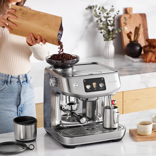 Silver espresso machine and grinder with a person pouring coffee beans into the grinder hopper with a modern looking kitchen background.