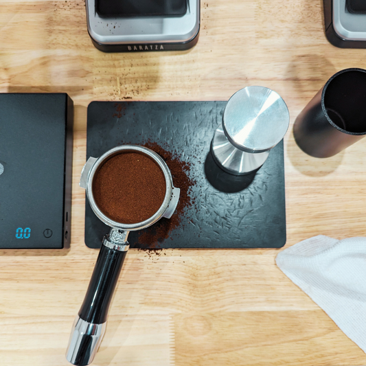 A portafilter with ground coffee in it, sitting on a tamping mat next to a tamper and coffee scale. 