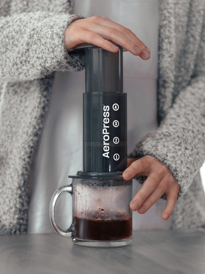Person using an Aeropress coffee maker with a glass mug on a gray surface.