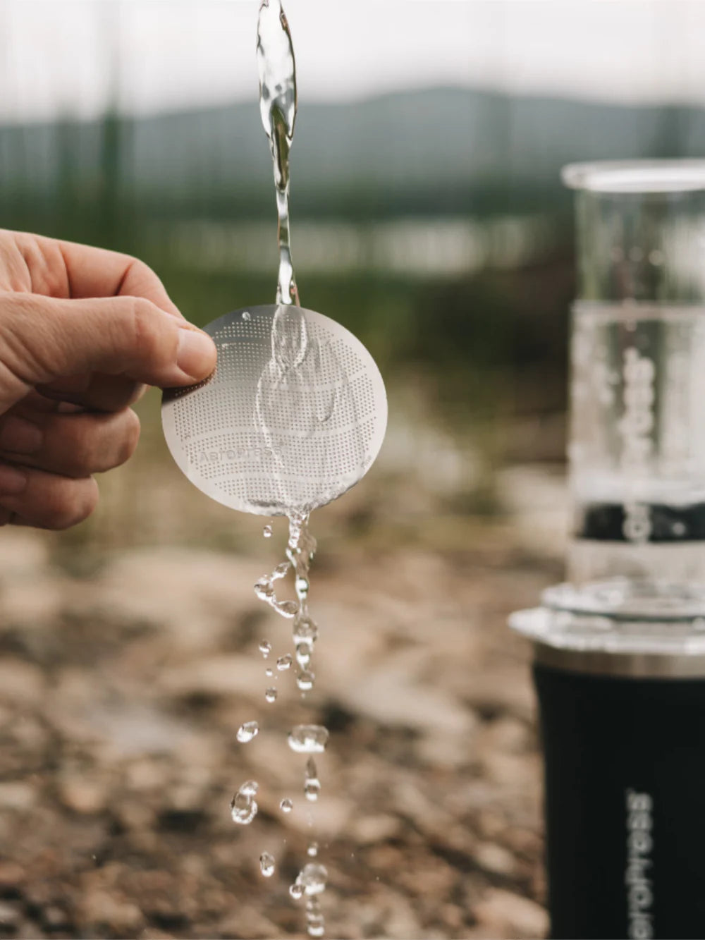 Hand holding a strainer with water dripping down with an outdoors background.