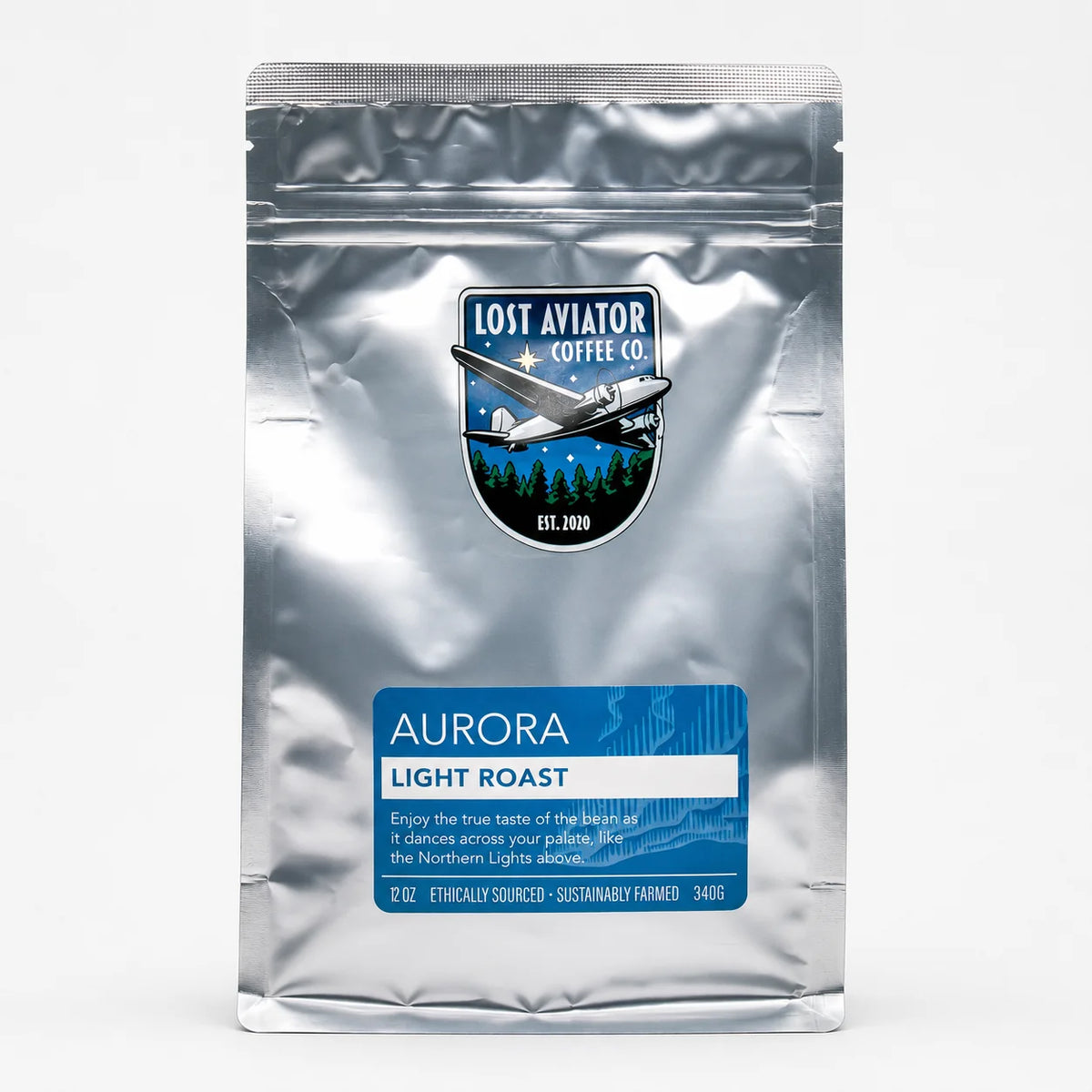 A silver stand-up pouch of Lost Aviator Coffee Co. Aurora Light Roast coffee, featuring a blue label and a logo with an airplane over a tree line, on a white background.