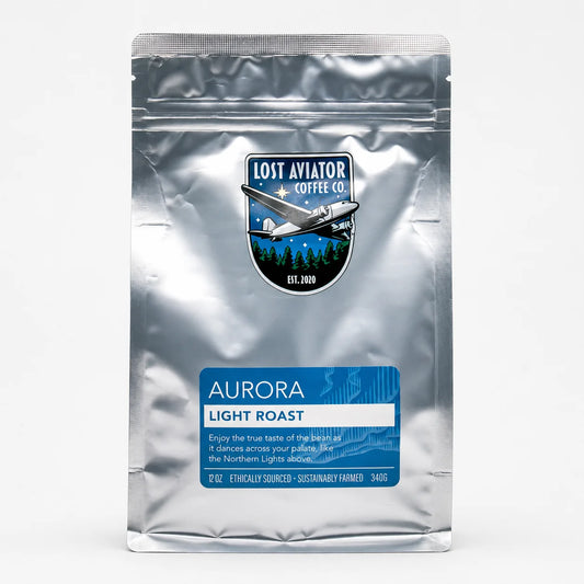A silver stand-up pouch of Lost Aviator Coffee Co. Aurora Light Roast coffee, featuring a blue label and a logo with an airplane over a tree line, on a white background.