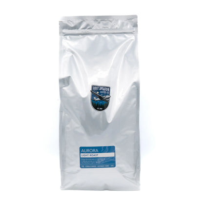 A silver foil bag Aurora Light Roast coffee, featuring a blue and white label near the bottom and a small shield logo in the center, set against a plain white background.