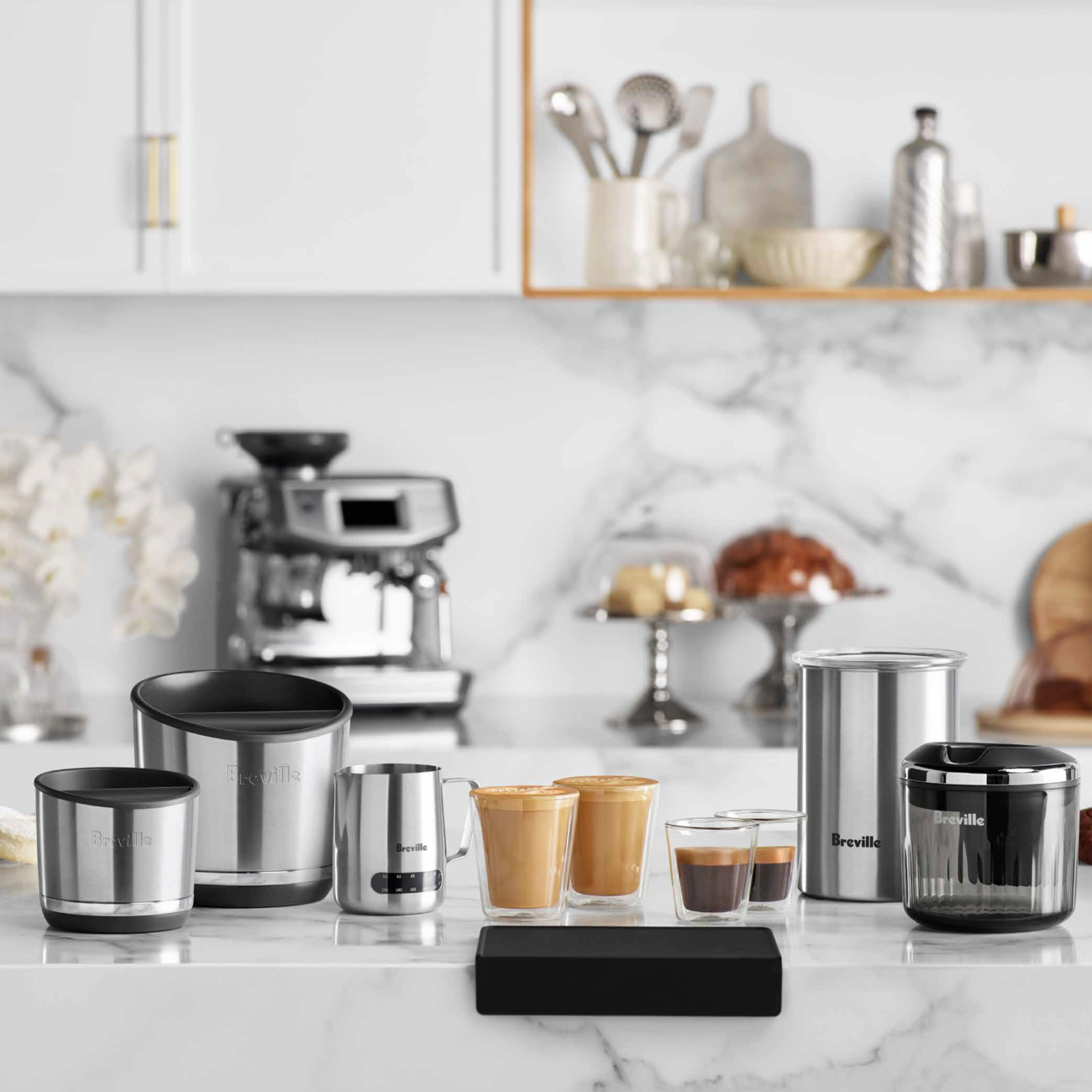 Collection of stainless steel kitchenware on a marble countertop with a coffee machine in the background.