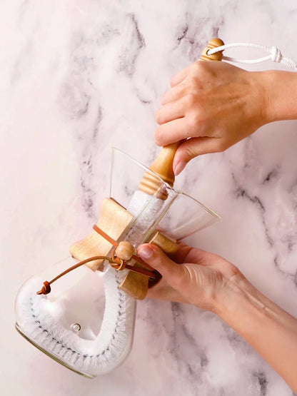 Person using a large cleaning brush on a glass coffeemaker against a marble background