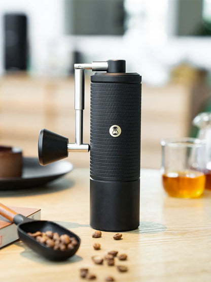 Black manual coffee grinder on a wooden table with coffee beans and a glass of coffee.