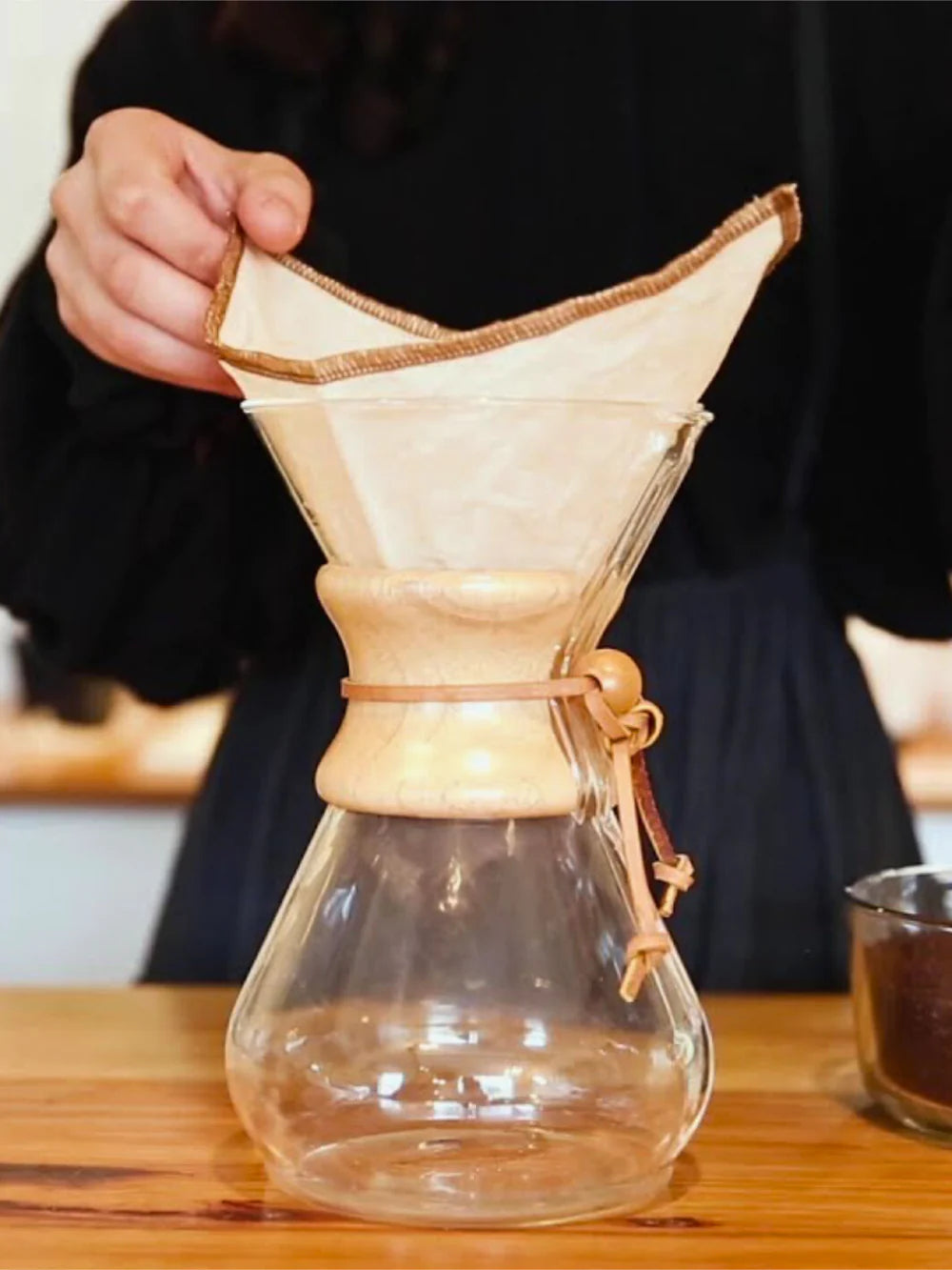 Person using a clear glass coffee maker with a wooden collar on a wooden table.