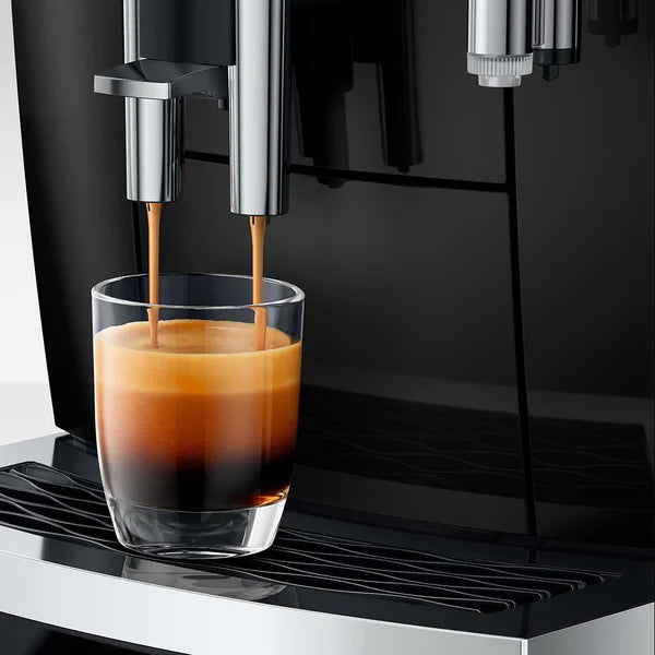 A close-up of a coffee machine dispensing espresso into a clear glass cup, set against a dark machine body and drip tray.