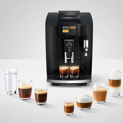 A black Jura coffee machine with two espresso shots and several layered coffee drinks and a glass of water arranged on a white surface against a light gray background.