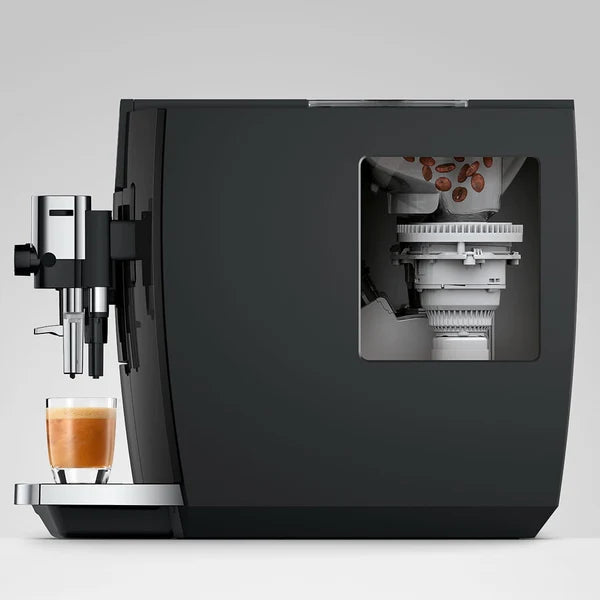 A modern dark gray coffee machine with a transparent window showing internal components, alongside a glass of espresso on a light gray background.