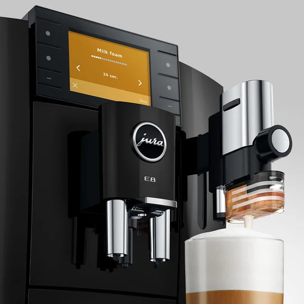 A close-up of a black Jura coffee machine with a digital touch screen, milk foam dispenser, and stainless steel components, shown dispensing milk foam into a cup against a light gray background.