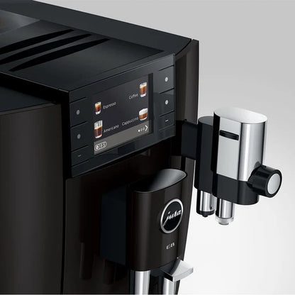 Close-up of a black Jura coffee machine with a digital control panel, drip tray, and a stainless steel steam wand attachment on a light gray background.