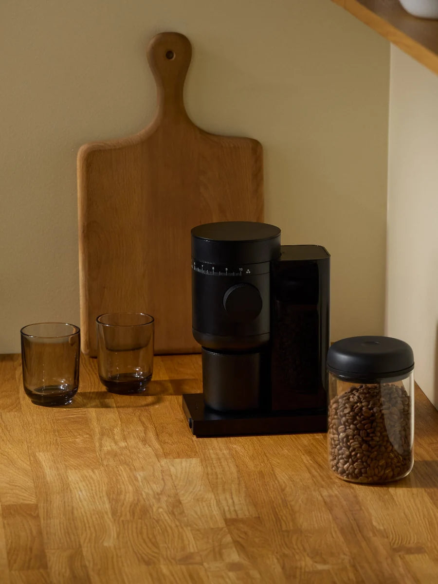 A black countertop coffee grinder with a matching bean jar and two dark glasses on a wooden surface, set against a beige wall with a wooden cutting board behind it.