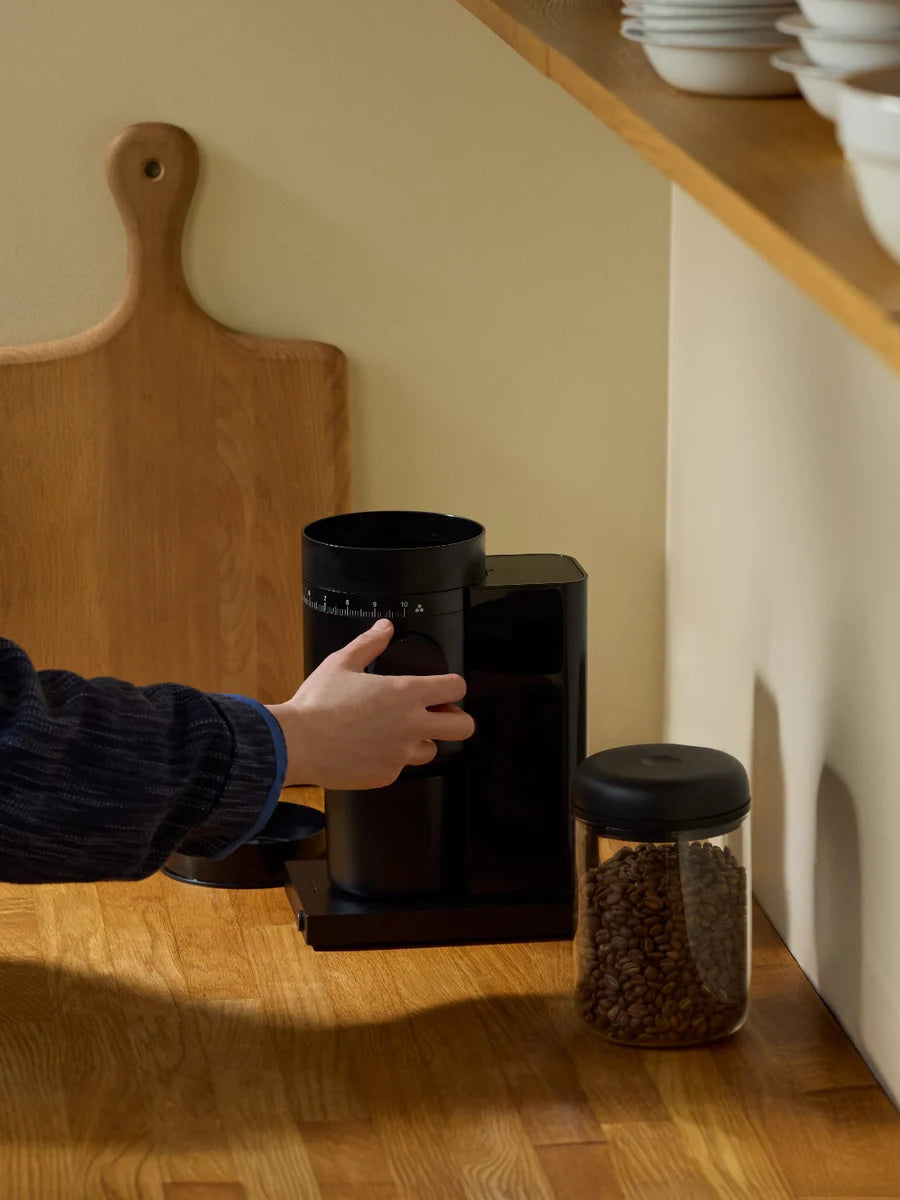Person using a coffee grinder on a wooden countertop with a jar of coffee beans nearby.