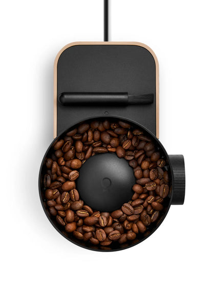 A top-down view of a modern coffee grinder with a matte black body, light wood-grain edges, and a circular opening filled with roasted coffee beans, shown on a plain white background.