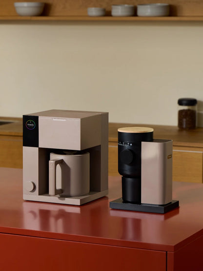 A modern coffee maker and a grinder sit side by side on a glossy red countertop in a minimalist kitchen setting with warm beige walls and wooden shelves.