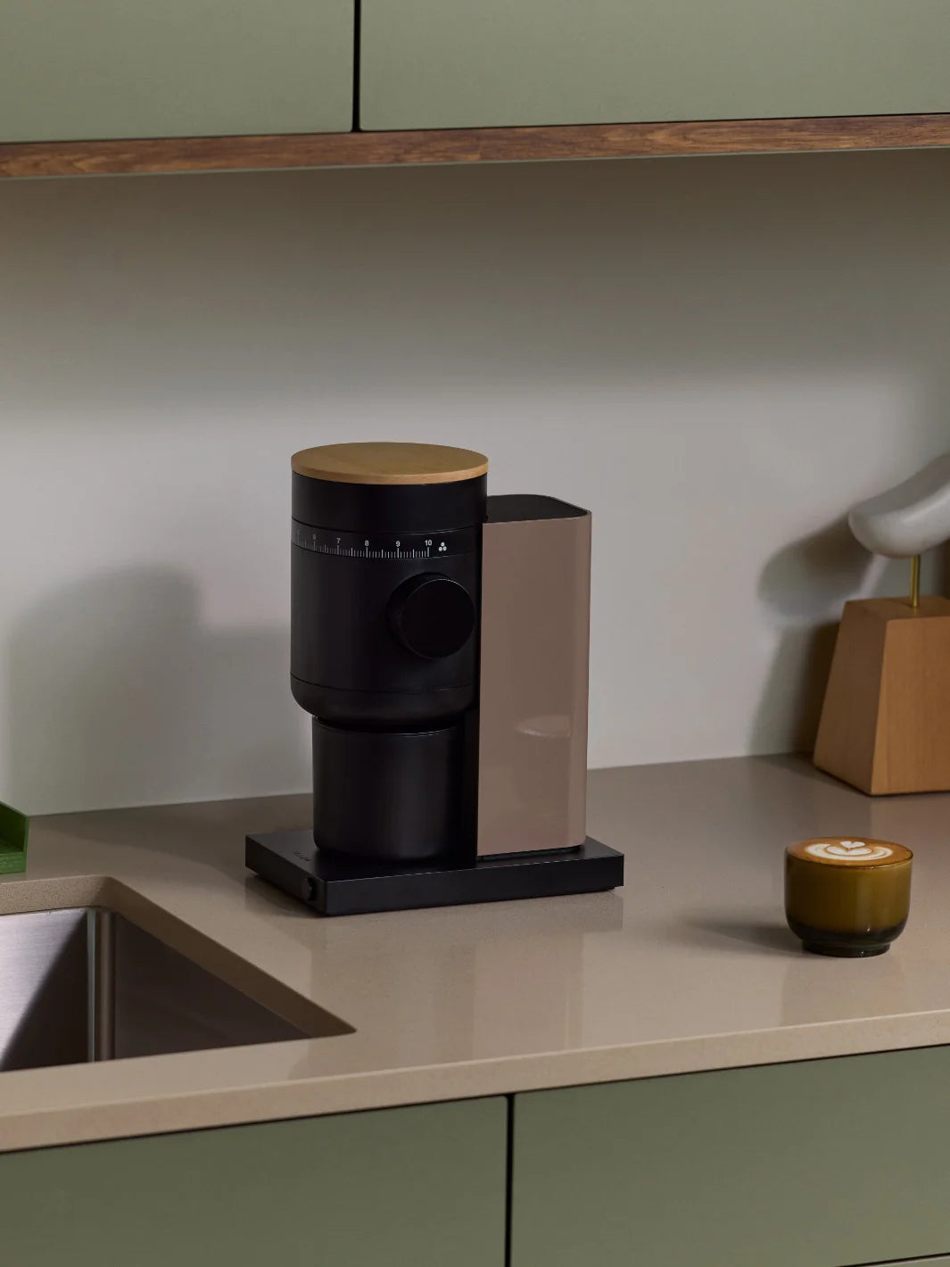 A modern coffee grinder with a matte black body, a light wood top, and a metallic bronze side panel, set on a beige kitchen countertop next to a sink.