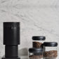 Black coffee grinder with glass Fellow Atmos canisters with coffee beans inside, on a marble surface