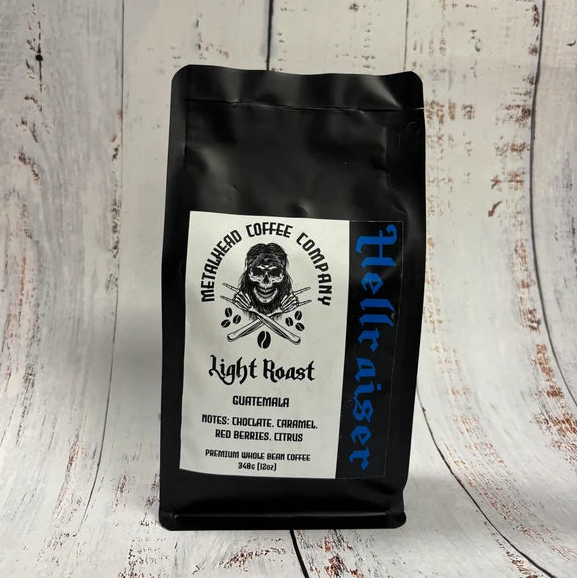 A black bag of Hellbitter Coffee Company Light Roast Guatemalan coffee stands upright on a whitewashed wooden surface. The bag features a white label with a skull graphic and text, and the brand name 'Hellbitter' printed vertically in blue on the right side.