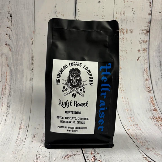 A black bag of Hellbitter Coffee Company Light Roast Guatemalan coffee stands upright on a whitewashed wooden surface. The bag features a white label with a skull graphic and text, and the brand name 'Hellbitter' printed vertically in blue on the right side.