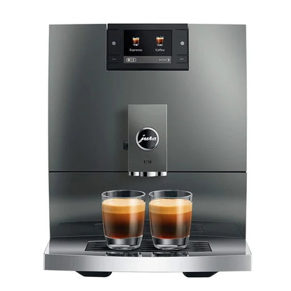 A sleek Jura C9 automatic espresso machine in dark gray stainless steel, shown with two freshly brewed espresso cups on its drip tray against a solid white background.