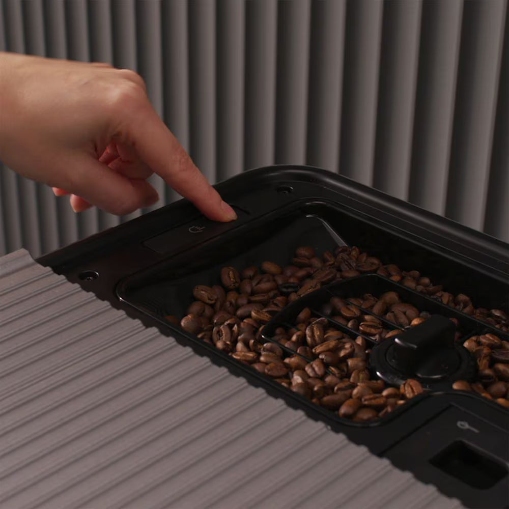 A close-up of a hand pointing toward a button on a black coffee bean hopper tray filled with coffee beans, set against a ribbed gray background.
