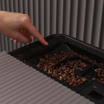 A close-up of a hand pointing toward a button on a black coffee bean hopper tray filled with coffee beans, set against a ribbed gray background.