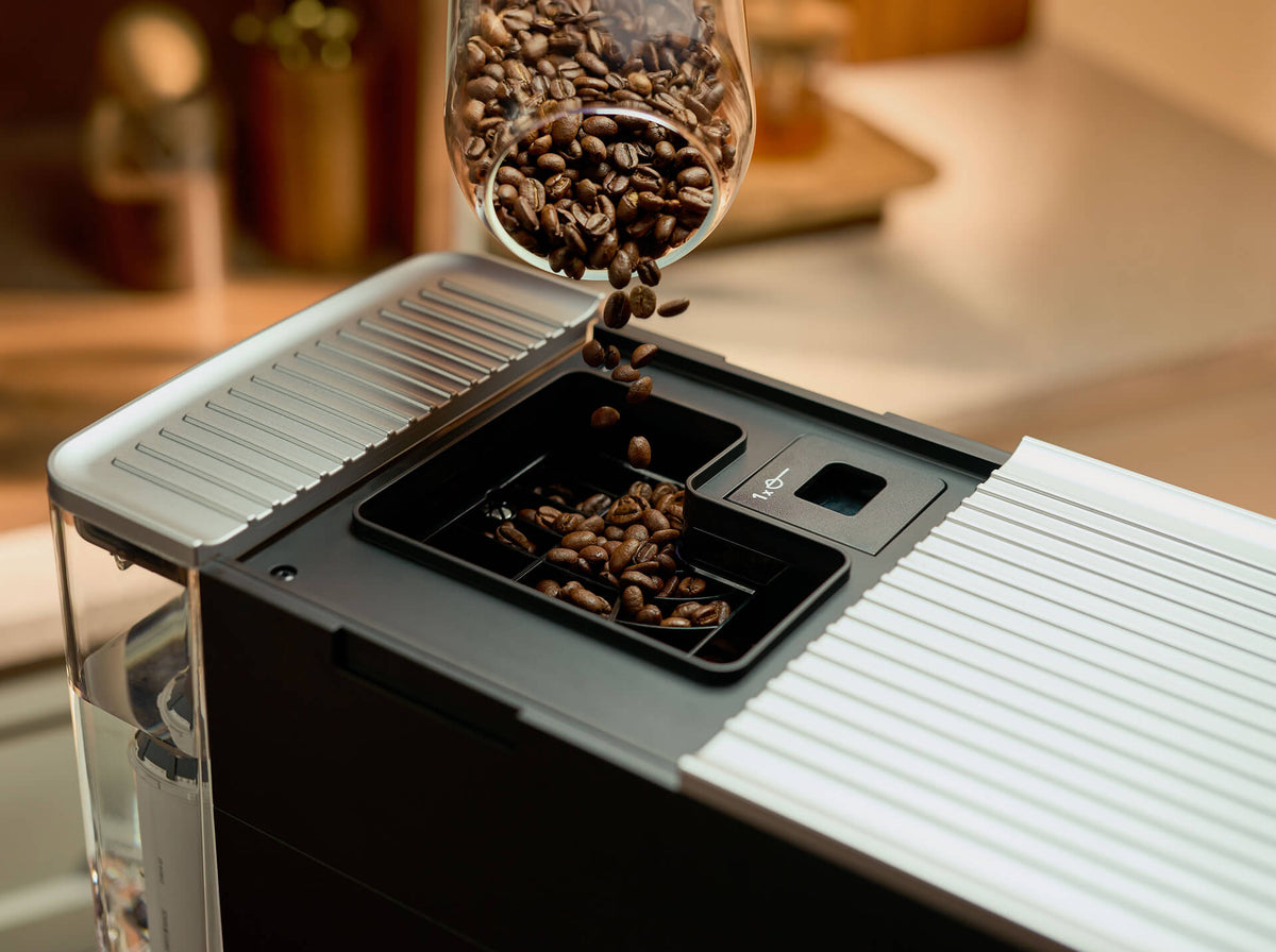 Coffee beans pouring from a glass container into a black and silver coffee machine, with a warm blurred kitchen background.