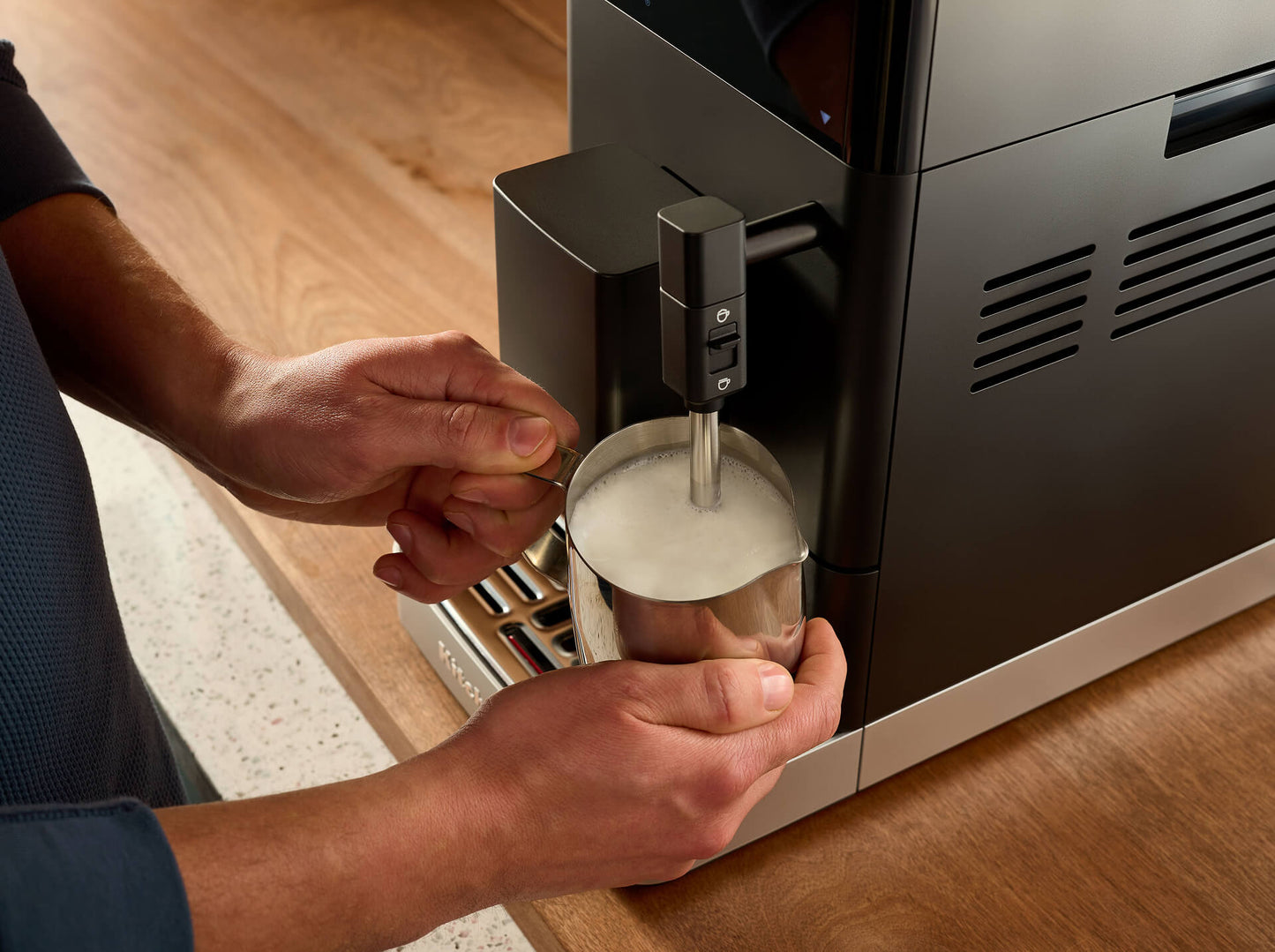A close-up of a person using a modern black and stainless steel espresso machine to steam milk in a metal pitcher, set on a wooden countertop.
