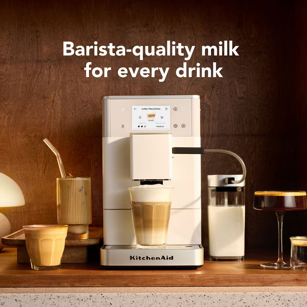 A KitchenAid barista-style milk dispenser machine sits on a countertop with a glass of milk and layered coffee drinks around it. Large text above reads 'Barista-quality milk for every drink.