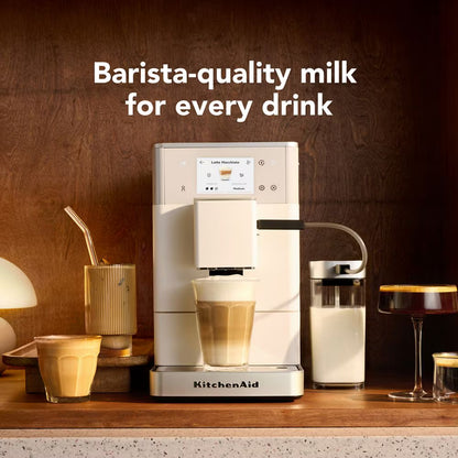 A KitchenAid barista-style milk dispenser machine sits on a countertop with a glass of milk and layered coffee drinks around it. Large text above reads 'Barista-quality milk for every drink.