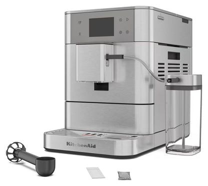 KitchenAid stainless steel automatic espresso machine with a digital display screen, shown with a clear water tank, black milk frother, and two small accessory packets on a white background.