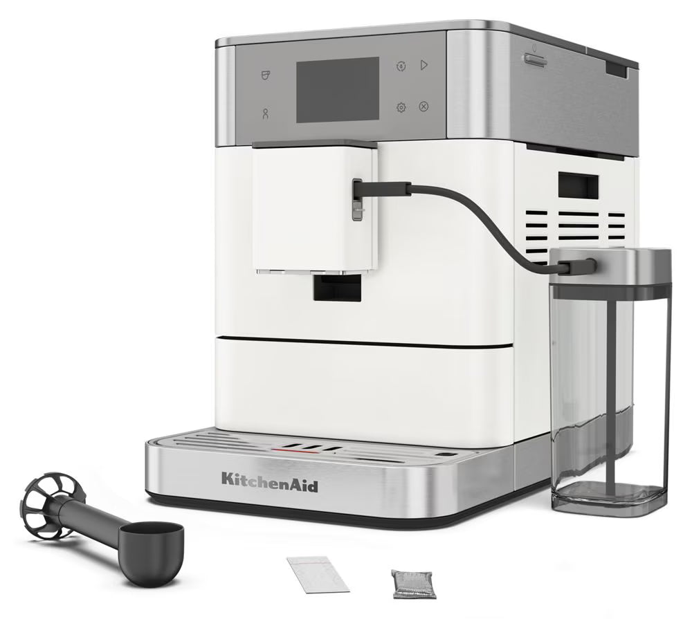 KitchenAid fully automatic espresso machine in white and stainless steel, shown with a glass water reservoir, black milk pitcher, and two small accessory packets on a clean white background.