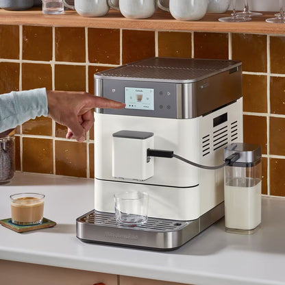 A countertop coffee machine with a cream body and stainless steel top, shown in a kitchen setting with a person's hand pointing at the control screen. A glass cup sits on the drip tray, and a milk container is connected on the right.