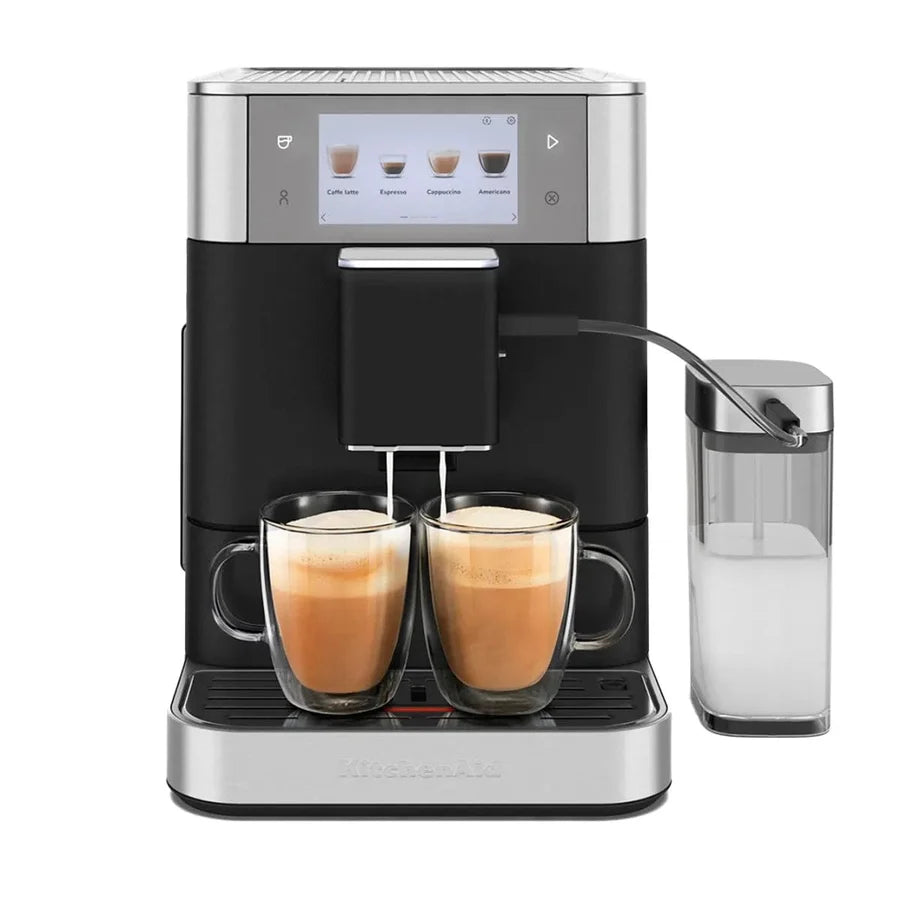 A KitchenAid espresso machine in black and silver, shown with two glass mugs of cappuccino on its drip tray and a milk container on the right, against a solid white background.