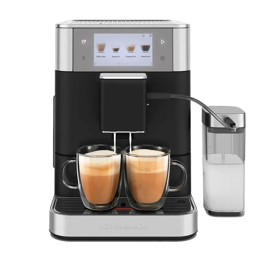 A KitchenAid espresso machine in black and silver, shown with two glass mugs of cappuccino on its drip tray and a milk container on the right, against a solid white background.