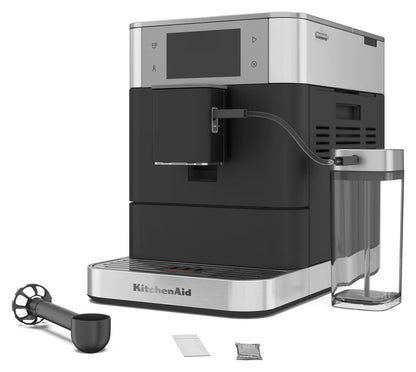 KitchenAid full-automatic espresso machine in black and stainless steel, shown with a clear water reservoir, black milk pitcher, and two sachets on a white background.
