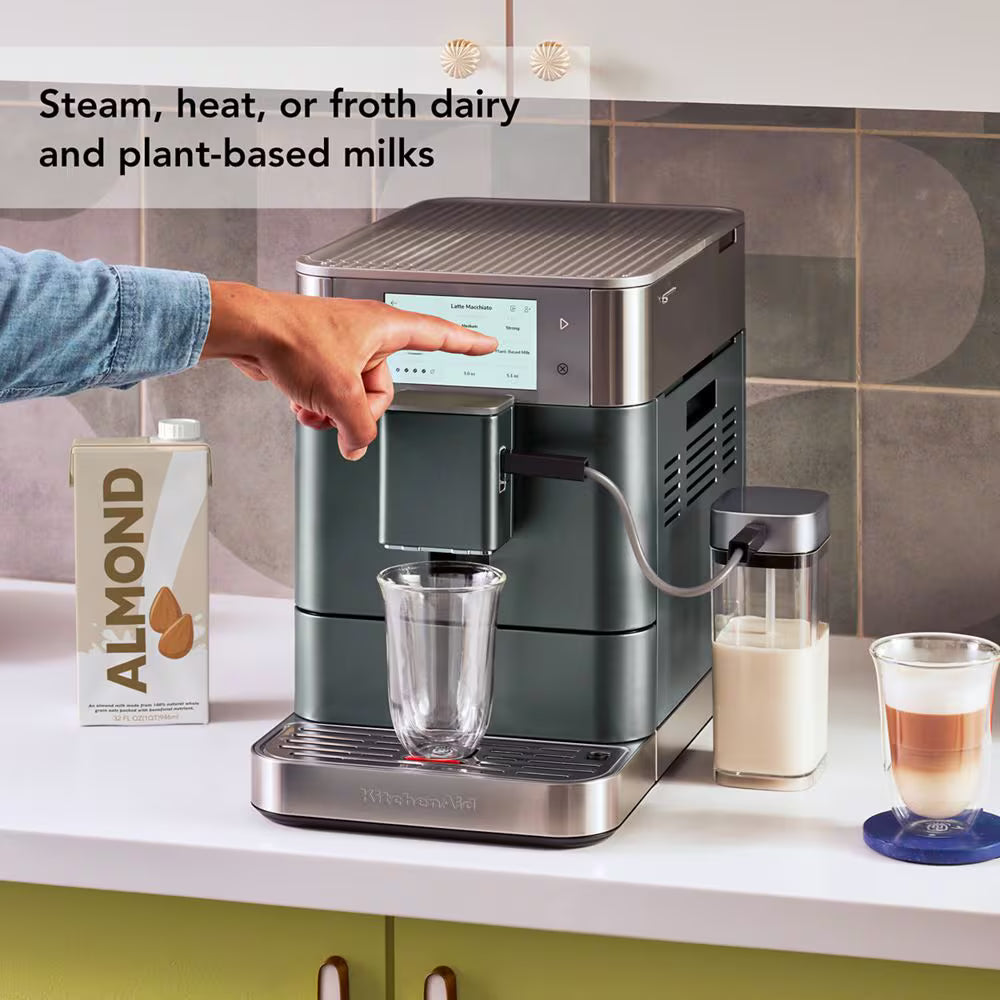 A KitchenAid coffee machine in dark green and stainless steel sits on a kitchen counter with a person's hand pressing the button to dispense milk. A carton of almond milk, a small milk bottle, and a latte are nearby, with text overlay reading 'Steam, heat, or froth dairy and plant-based milks'.