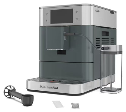 KitchenAid coffee machine in dark gray and stainless steel, shown with a clear water tank, black milk pitcher, and two small accessories on a white background.
