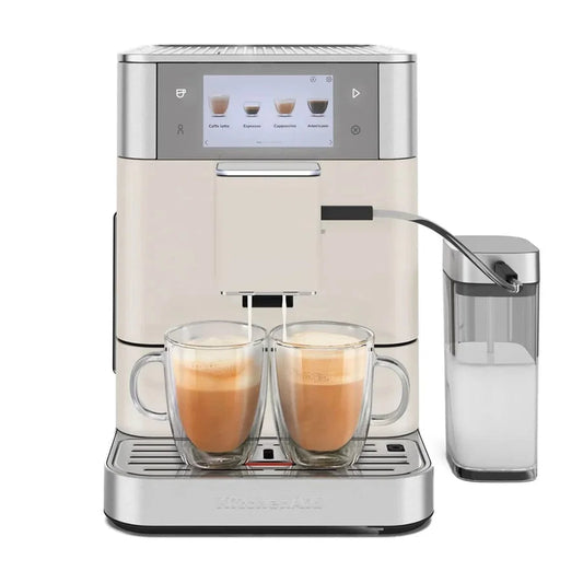 KitchenAid fully automatic espresso machine in cream and stainless steel, shown with two double-walled glass mugs of cappuccino and a milk carafe container on a white background.