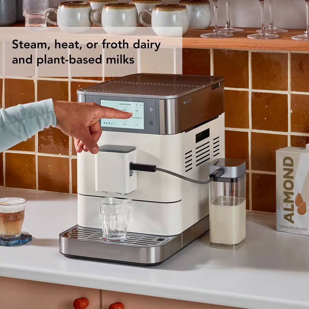 A KitchenAid coffee machine in a cream and stainless steel finish, shown on a kitchen counter with a hand pointing at the touch screen. A glass cup is under the spout, and a milk container is connected on the side.