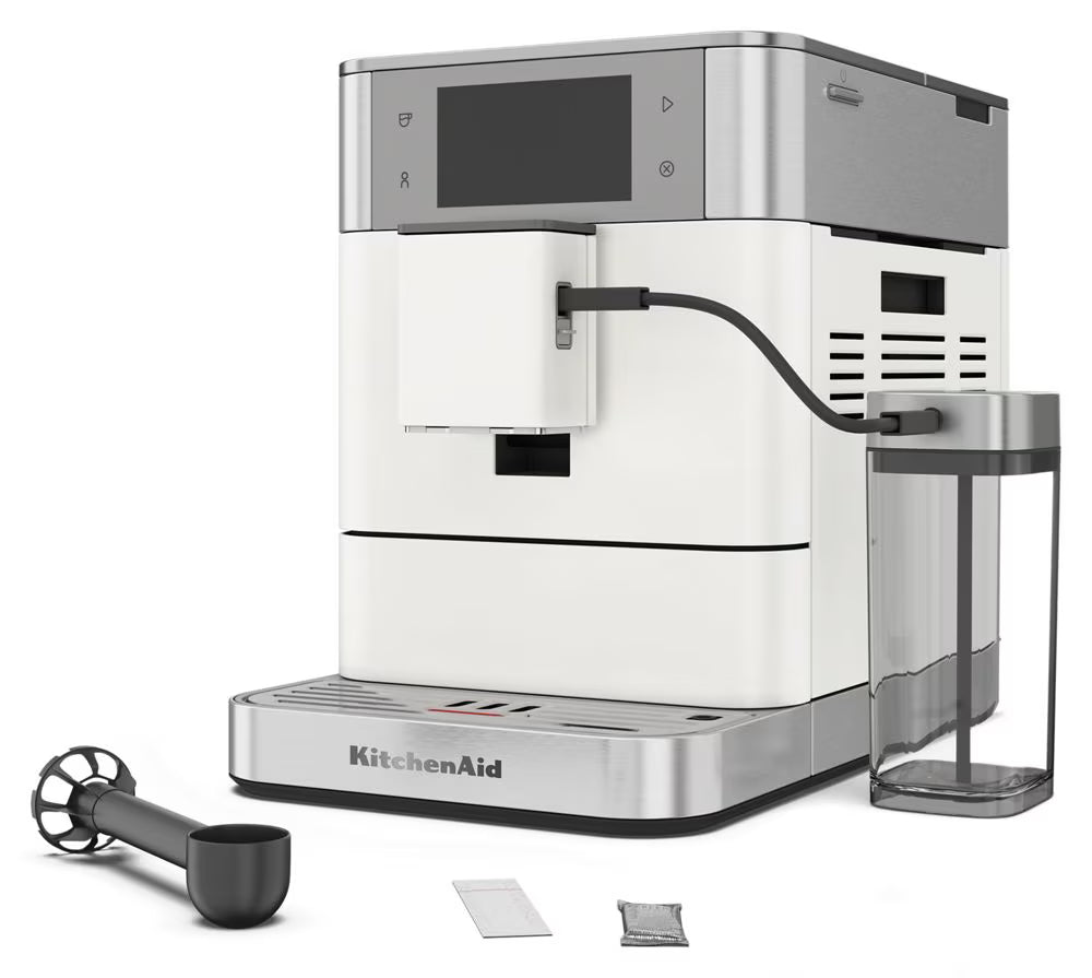 KitchenAid full-automated espresso machine in white and stainless steel, shown with a clear water reservoir, black milk pitcher, and small accessory packets on a white background.