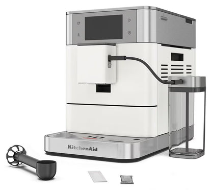 KitchenAid full-automated espresso machine in white and stainless steel, shown with a clear water reservoir, black milk pitcher, and small accessory packets on a white background.
