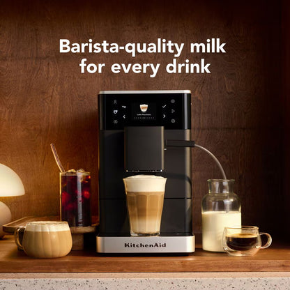 A KitchenAid coffee machine on a wooden countertop, shown dispensing milk into a latte and drawing cream from a milk bottle, with the text 'Barista-quality milk for every drink' above it.