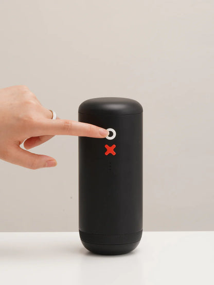 A matte black cylindrical portable coffee grinder with a white power button and a red X stop button, shown with a hand pointing at the button against a neutral beige background.